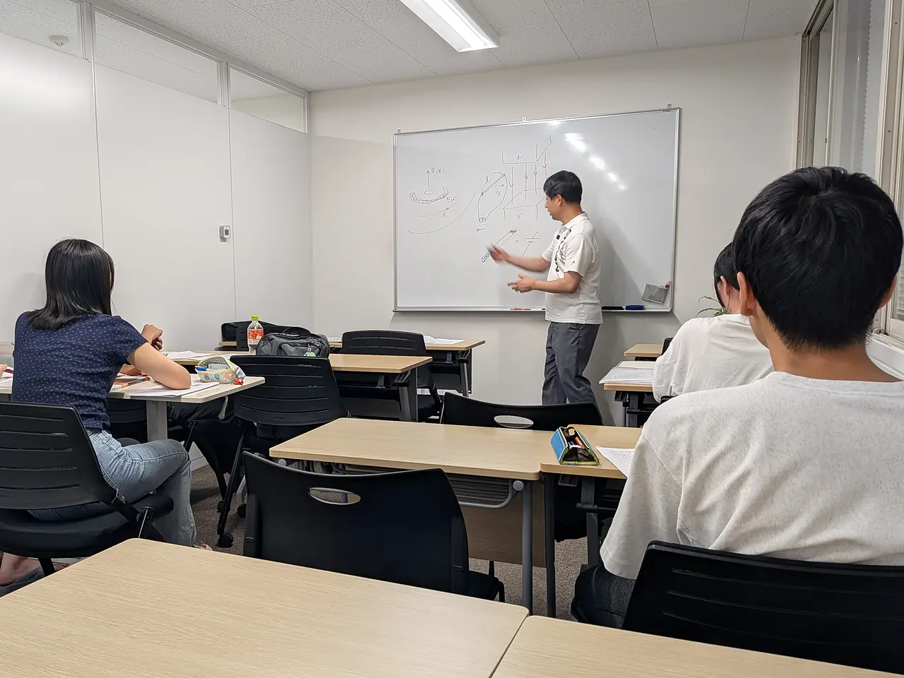 授業中の少人数教室。講師がホワイトボードに図を描きながら説明し、塾生が席から見ている